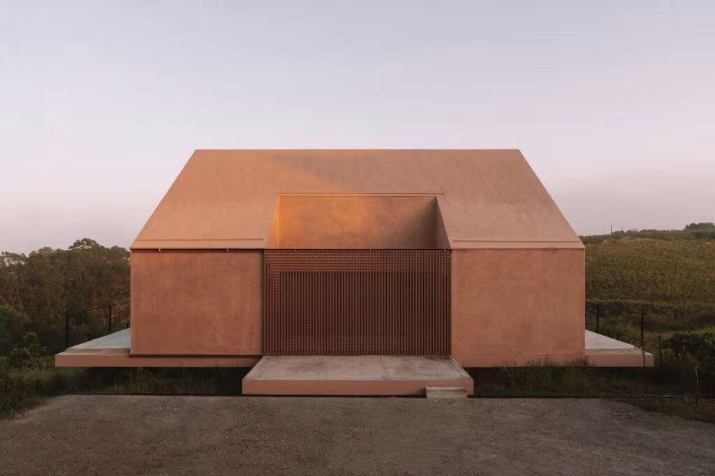 Casa Plaj, vivienda de tonos rojizos con cubierta a dos aguas y puerta de lamas sobre el prado