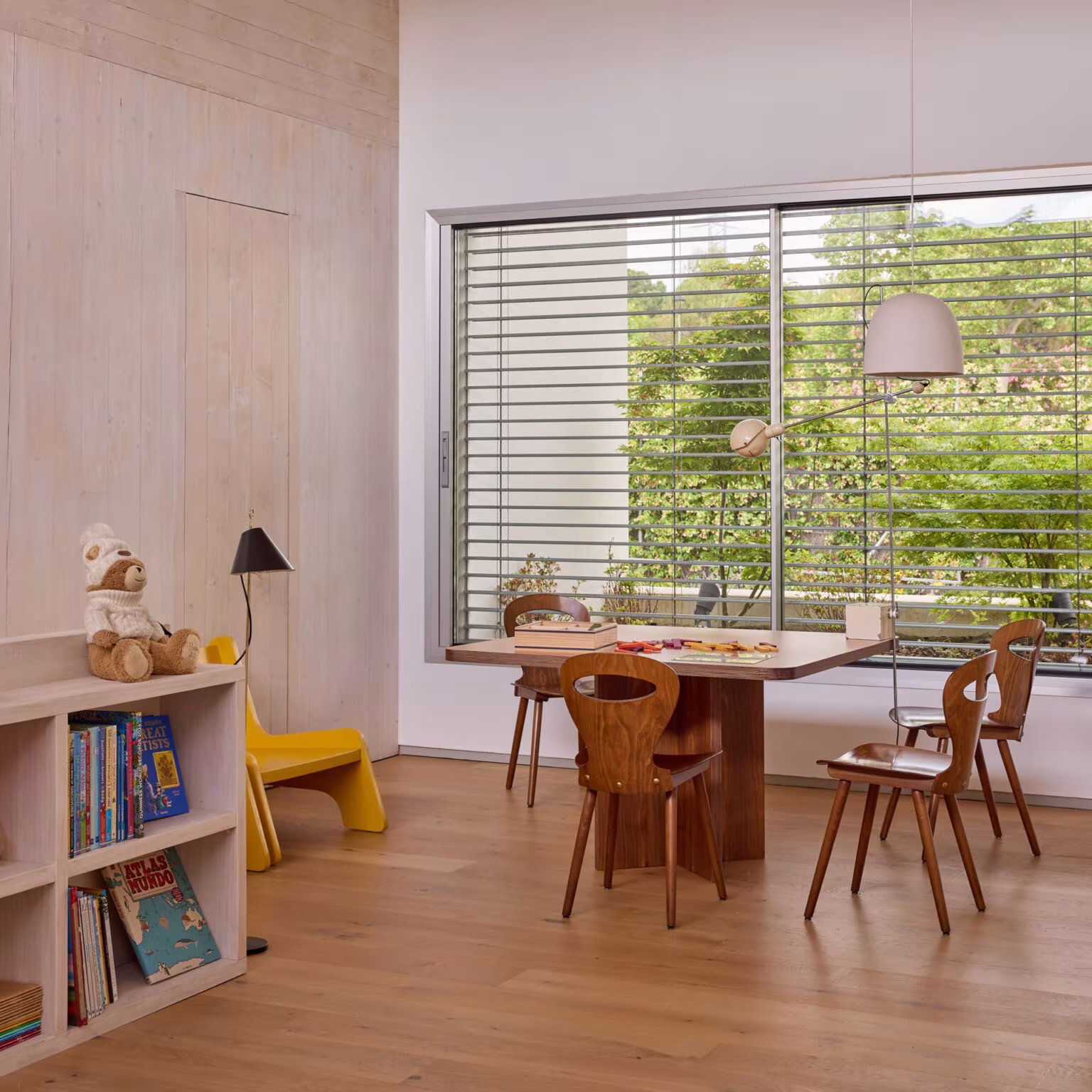 Sala de juegos infantil con mesa, sillas de madera y gran ventana con lamas al jardín en la Casa de los arces