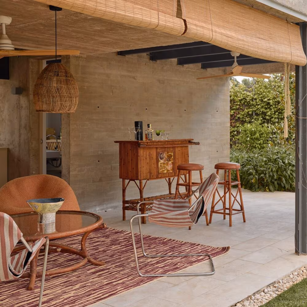 Porche con muros de hormigón, mueble bar de madera, butacas de rejilla y alfombra a rayas en el Proyecto AB de Ábaton