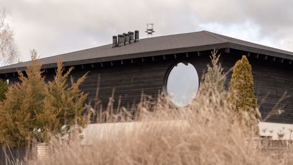 Fachada de madera quemada de la Dzen House con ventana circular bajo una cubierta de gran alero, entre hierbas altas