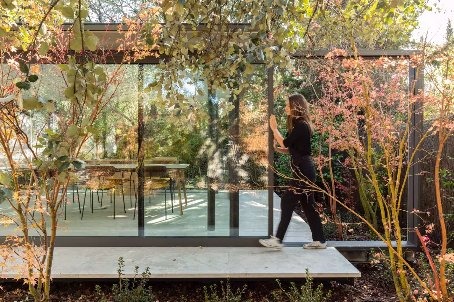 A woman walking next to a glass pavilion among autumn trees, in a project by Ábaton