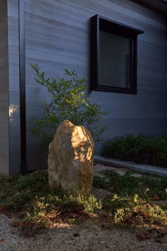Roca vertical como pieza escultórica ante una fachada de madera oscura con ventana, entre plantas bajas