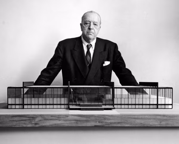 Mies van der Rohe in a dark suit behind a building model, in a black and white portrait