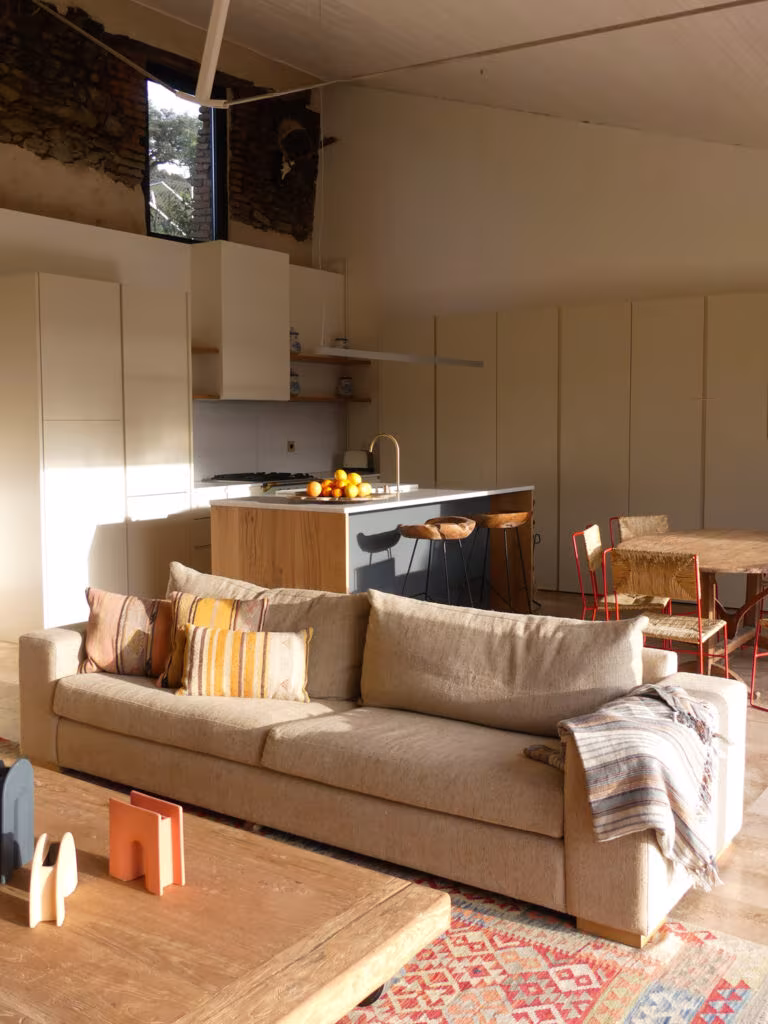 Living space with sofa, low table and timber-island kitchen beneath wooden beams in La Vera