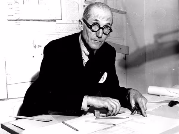 Black and white portrait of Le Corbusier in round glasses working over drawings