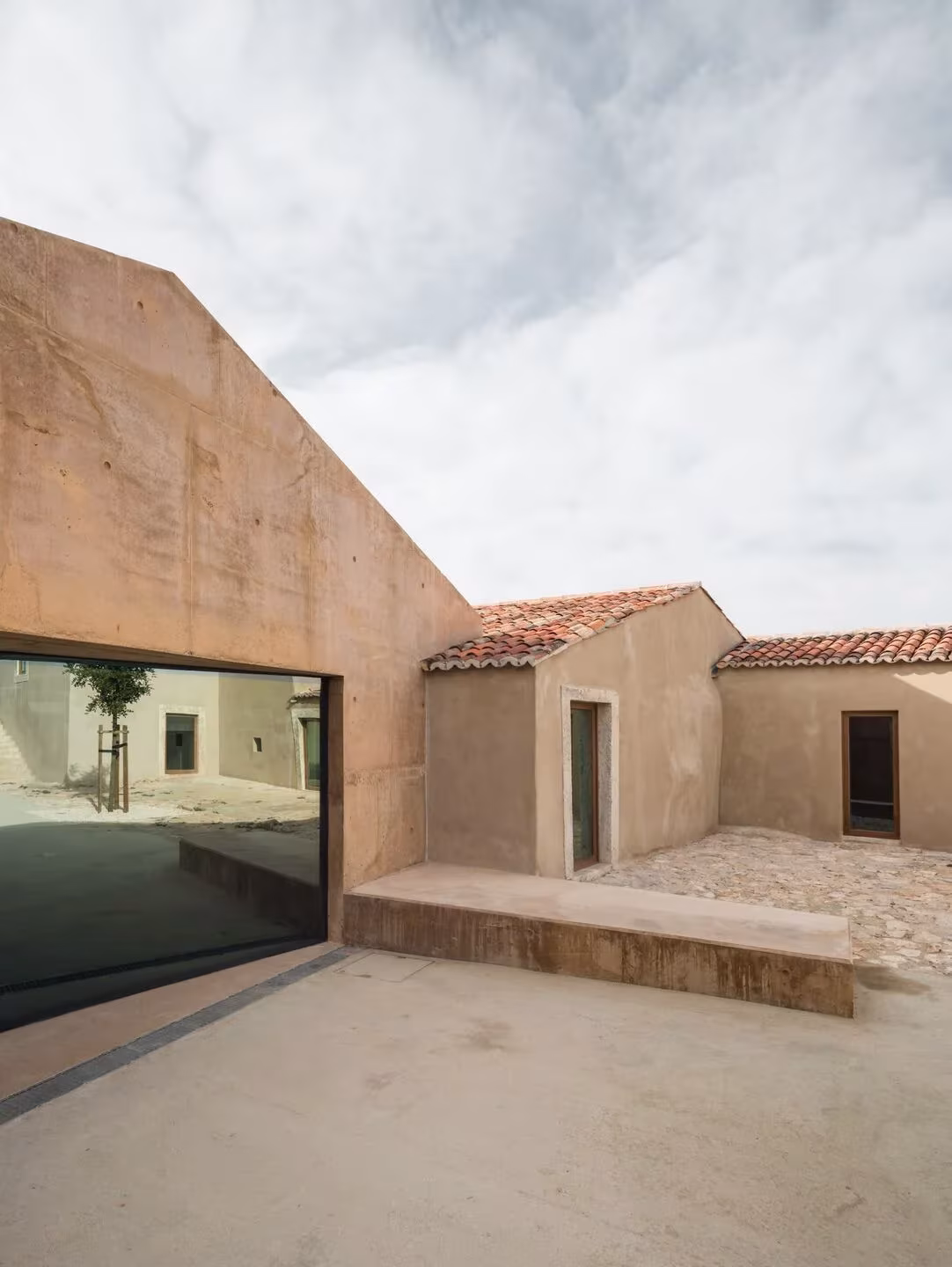 Patio del Casal Saloio con gran ventanal de vidrio oscuro en un volumen de hormigón rosado y banco de piedra