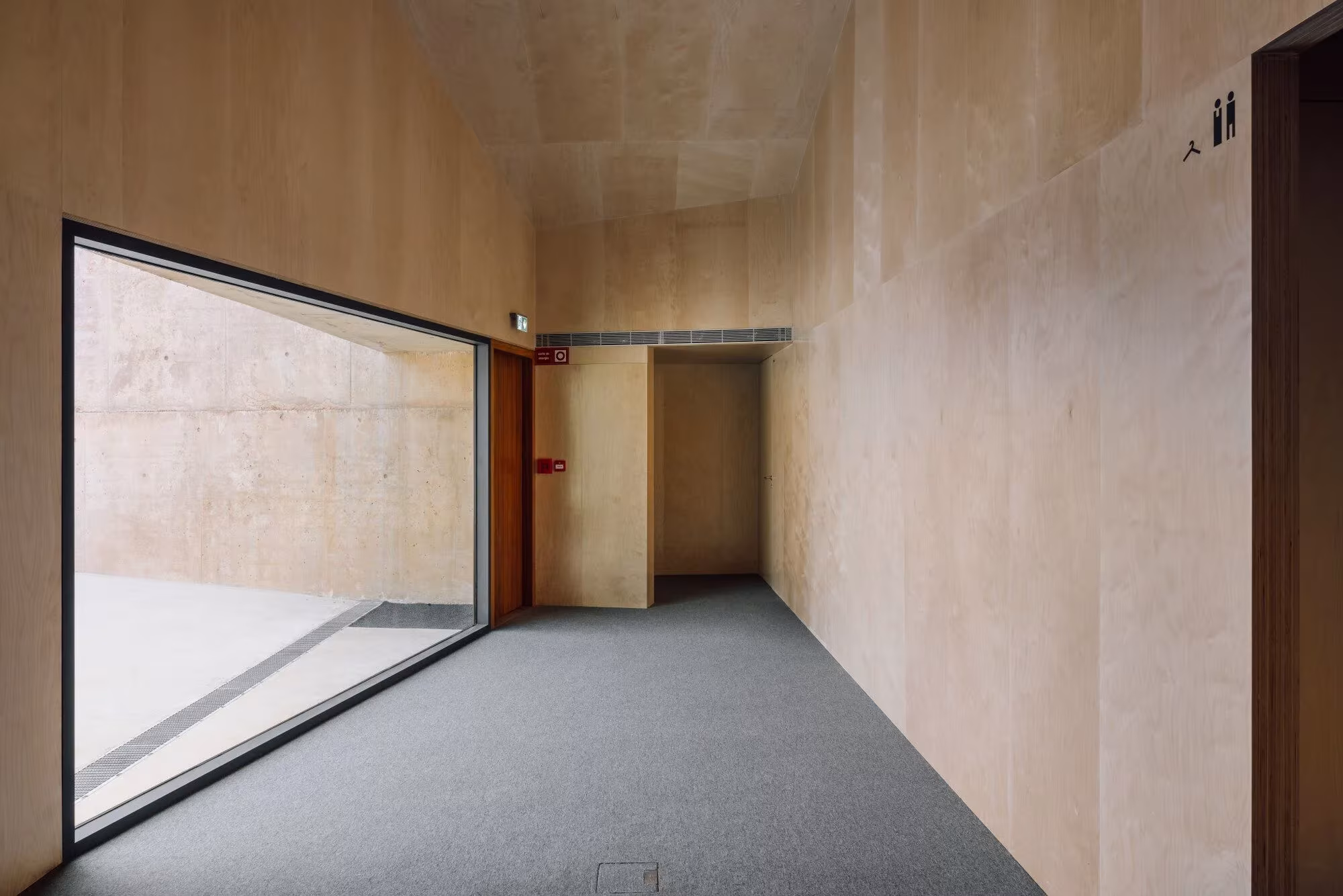 Pasillo de la sala del Casal Saloio con paramentos de madera clara y gran ventana a ras de suelo