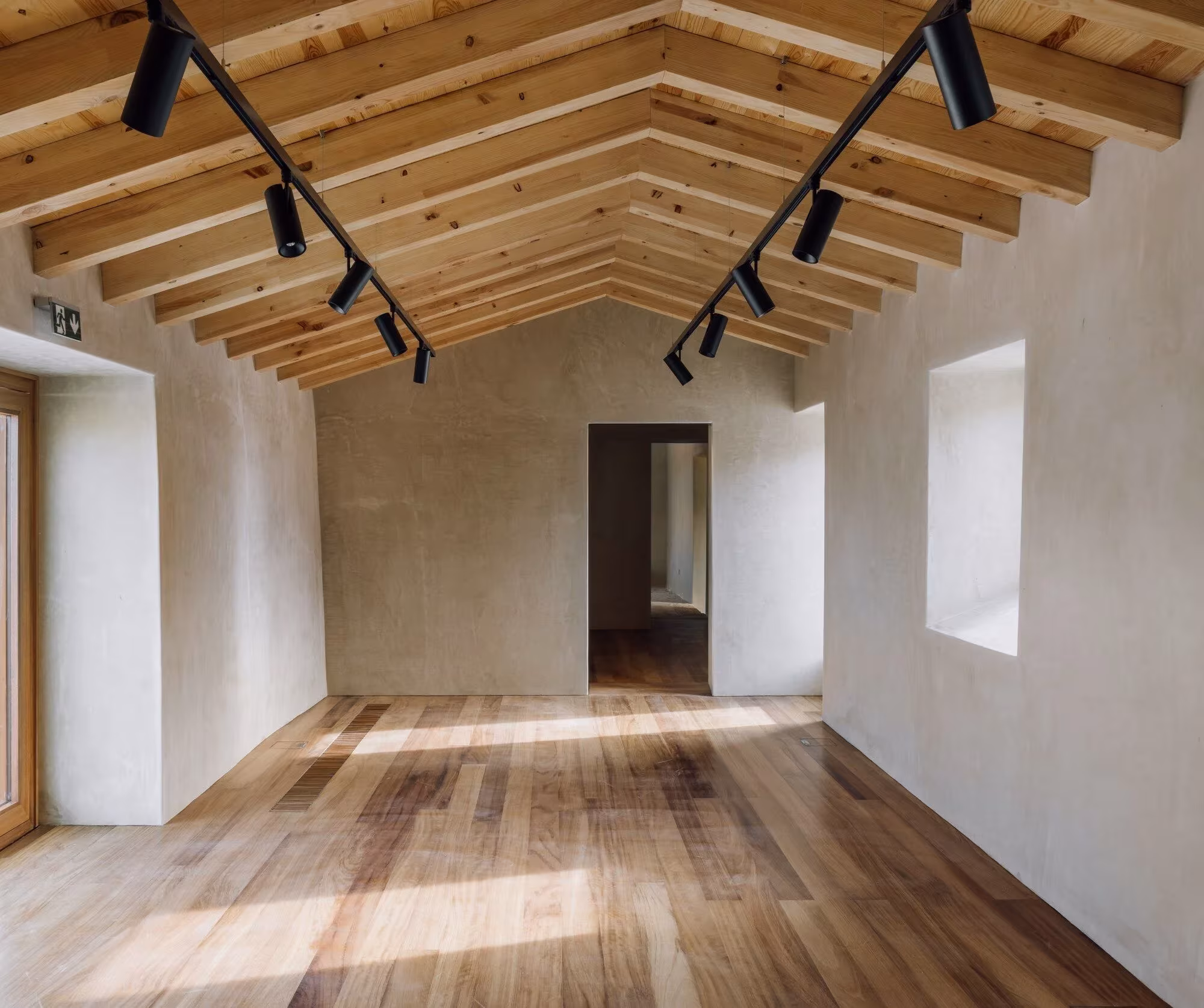 Sala de exposición bajo cubierta de madera a dos aguas con focos negros y suelo de tarima en el Casal Saloio
