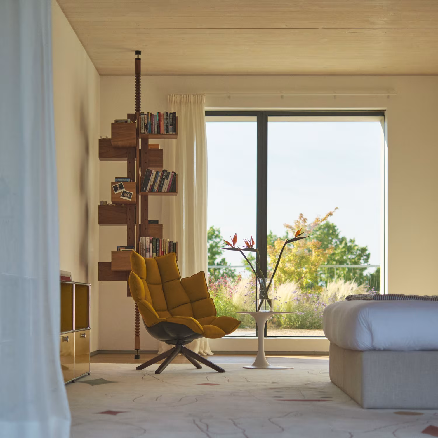 Dormitorio con librería giratoria de madera y butaca amarilla ante un gran ventanal sobre el jardín