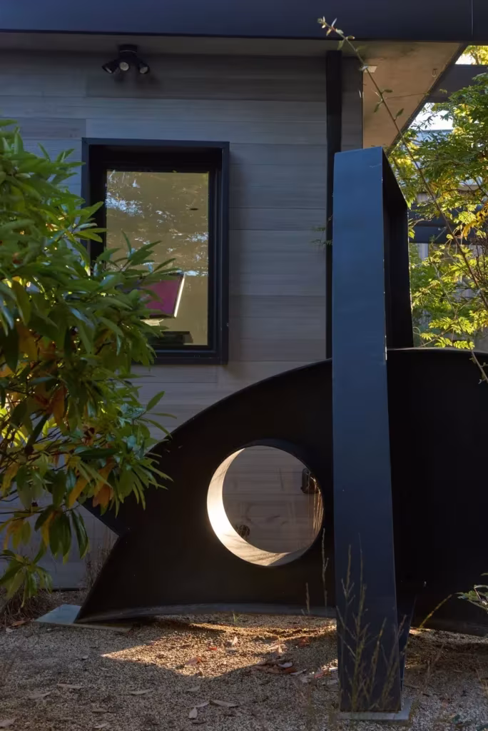 Escultura de acero negro con orificio circular frente a una fachada de madera y ventana de marco oscuro