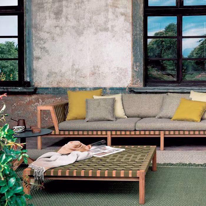 Sofá de exterior con cojines amarillos y mesa de centro de cuerda verde ante una pared desconchada con ventanales