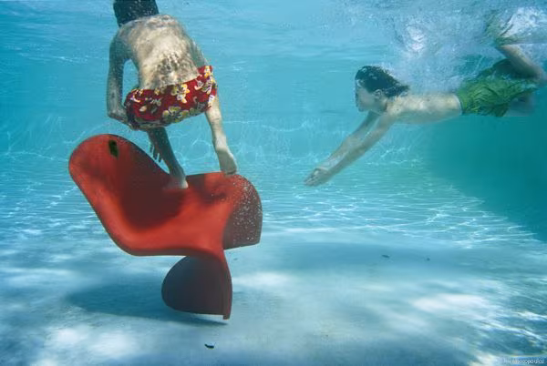 Two swimmers underwater beside a red Panton chair submerged in a pool