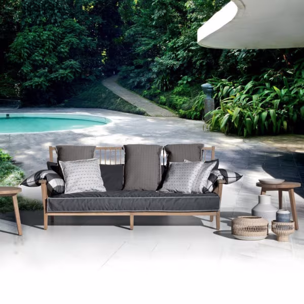 Teak outdoor sofa with grey cushions and woven side tables beside a pool among vegetation