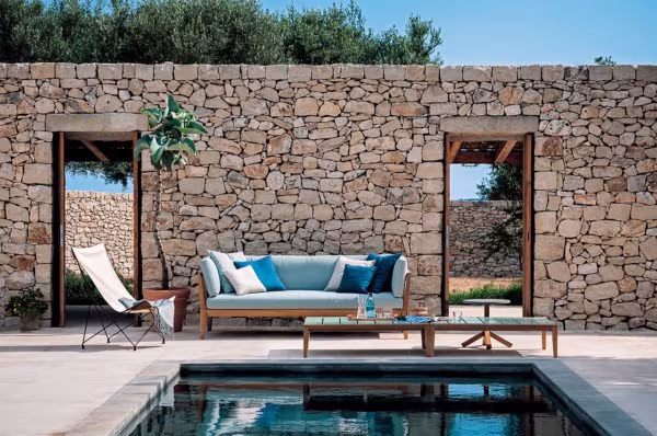 Outdoor sofa with blue cushions and a deck chair before a stone wall and a swimming pool