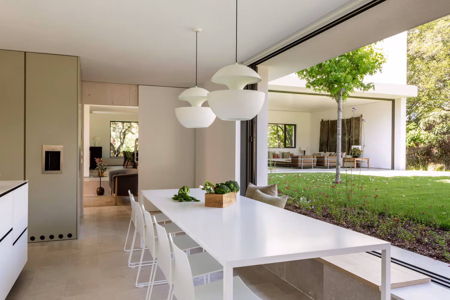 Mesa blanca alargada junto a la cocina con puertas correderas abiertas al césped en el Proyecto AF