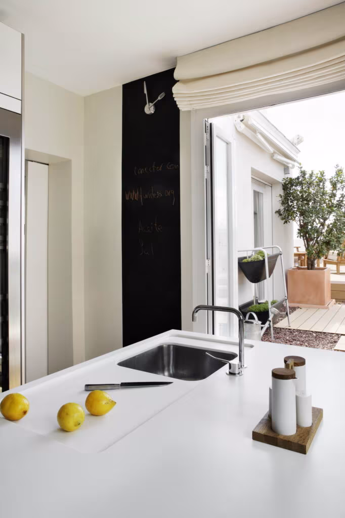 White kitchen island with sink opening onto a terrace with an olive tree and roman blind in the Rosales refurbishment by Ábaton