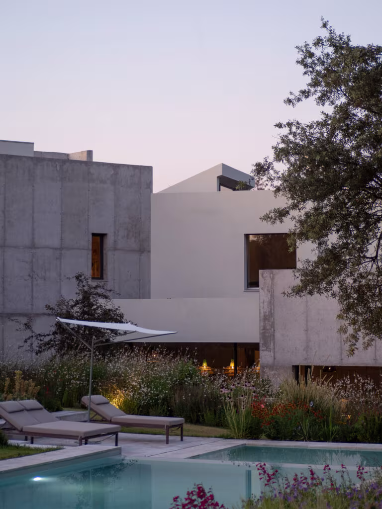 Volúmenes de hormigón y encalado de la Casa ÁRCO entre encinas y plantación junto a la piscina al crepúsculo