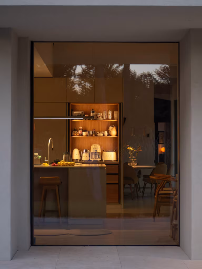 Cocina iluminada al atardecer vista a través de un gran hueco acristalado en la fachada de la Casa ÁRCO