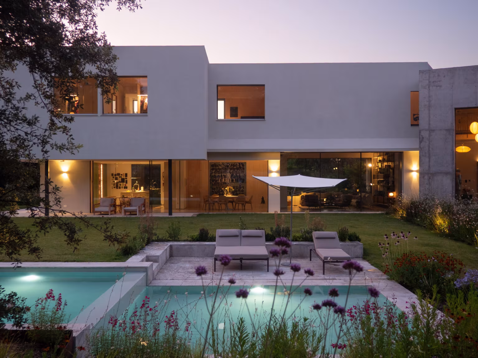 Casa ÁRCO al anochecer con las ventanas iluminadas sobre la piscina escalonada y las tumbonas