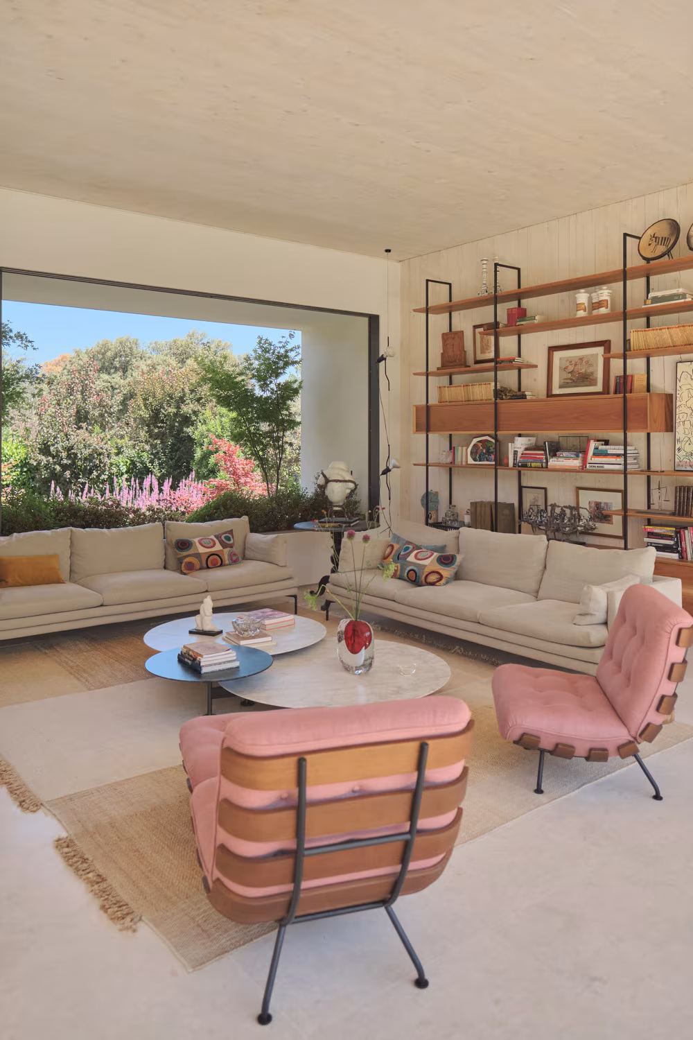 Sala de estar con butacas rosas, sofás y ventanal al jardín junto a la estantería de obras de la Casa ÁRCO