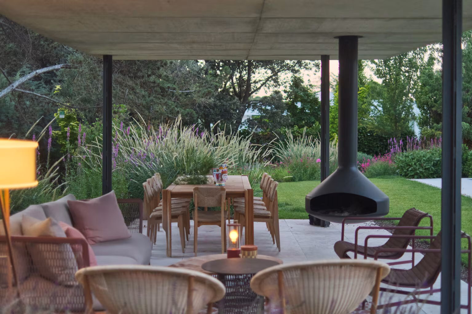 Porche de verano con estufa colgante, mesa comedor y butacas ante el jardín al atardecer en la Casa ÁRCO de Ábaton