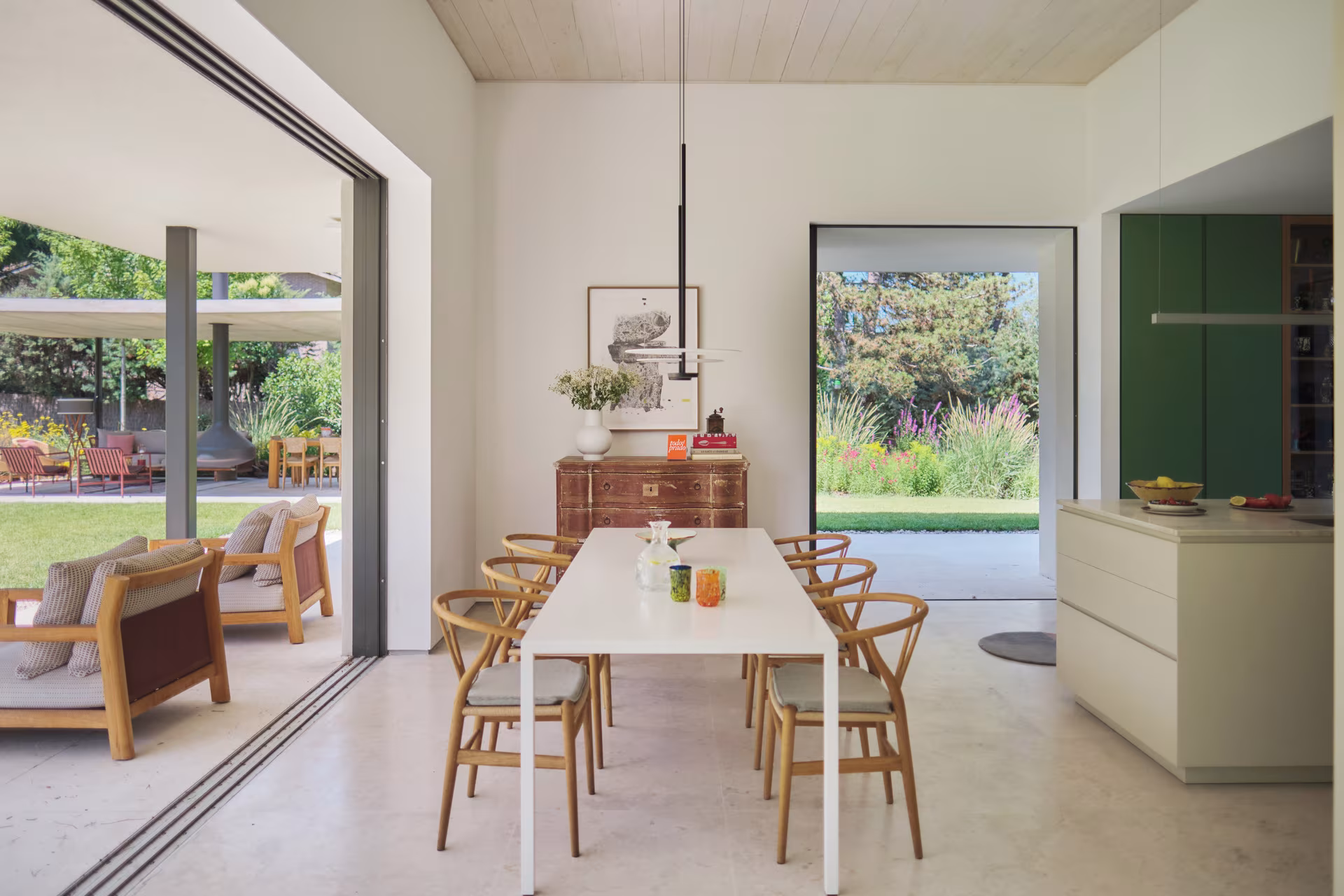 Cocina y comedor con mesa blanca alargada y sillas de madera abiertos al porche y al jardín en la Casa ÁRCO