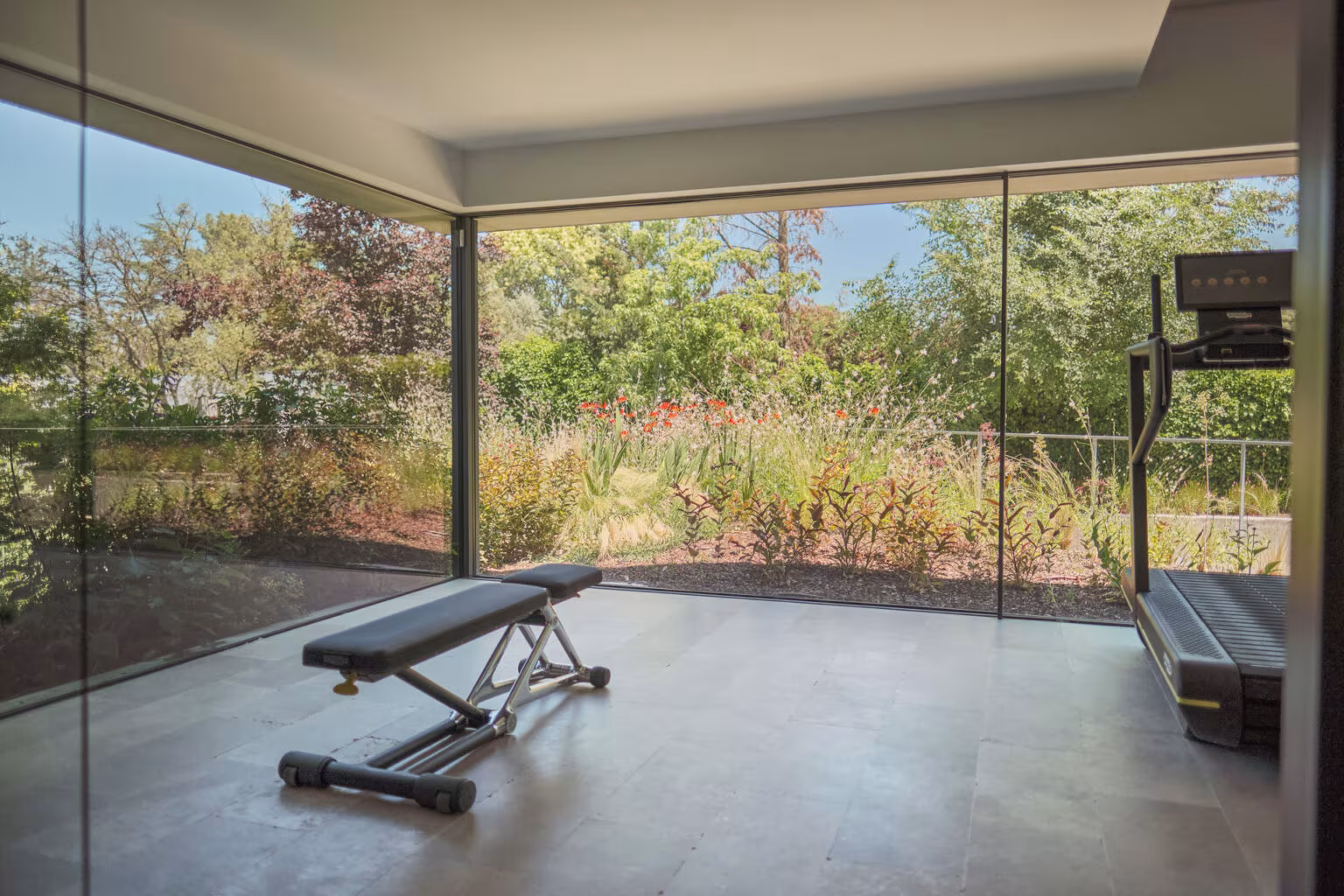 Sala de ejercicio acristalada con banco de pesas abierta a un jardín de plantación silvestre en la Casa ÁRCO de Ábaton
