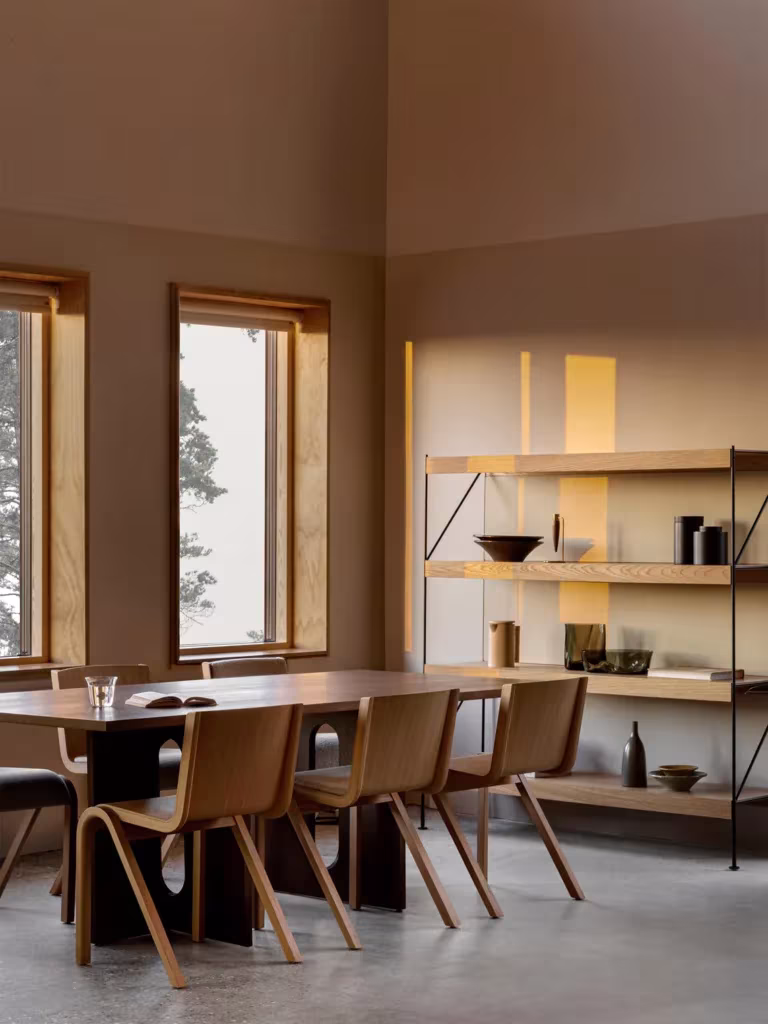 Comedor de madera con sillas de contrachapado, estantería abierta y ventanas con marco de madera en tonos cálidos