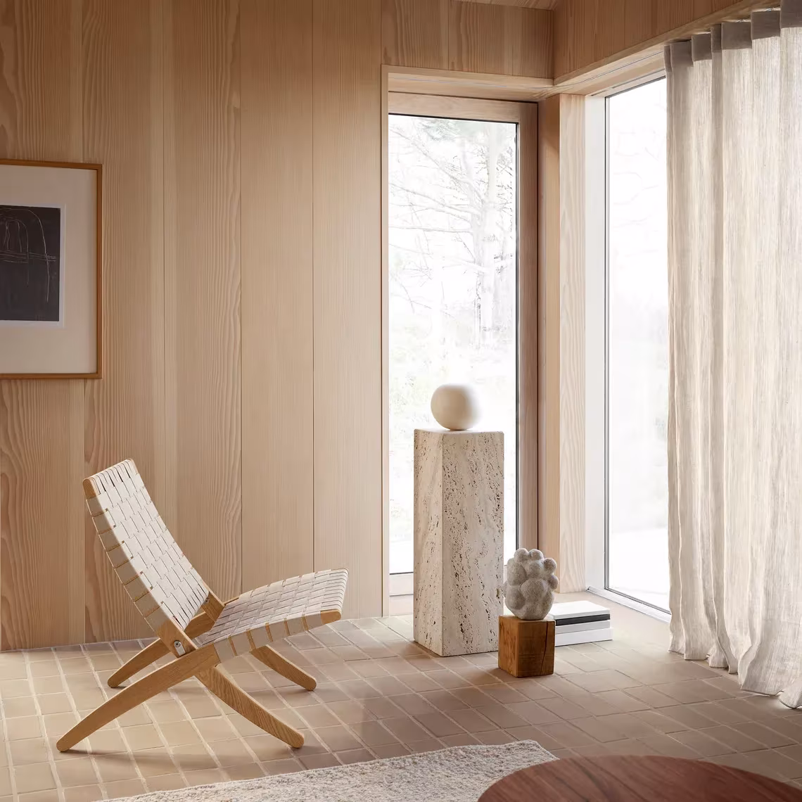 Butaca plegable de madera y cuerda junto a un pedestal de travertino en una sala revestida de madera clara