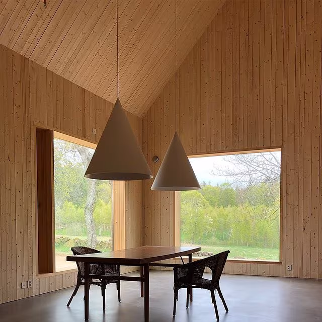 Comedor bajo cubierta de madera a dos aguas con lámparas cónicas, mesa y sillas de mimbre ante un gran ventanal