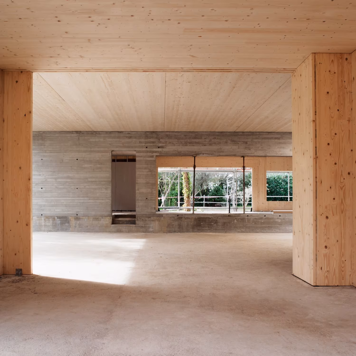 Sala amplia con estructura de madera laminada y muros de hormigón visto que se abre al jardín en una obra de Ábaton