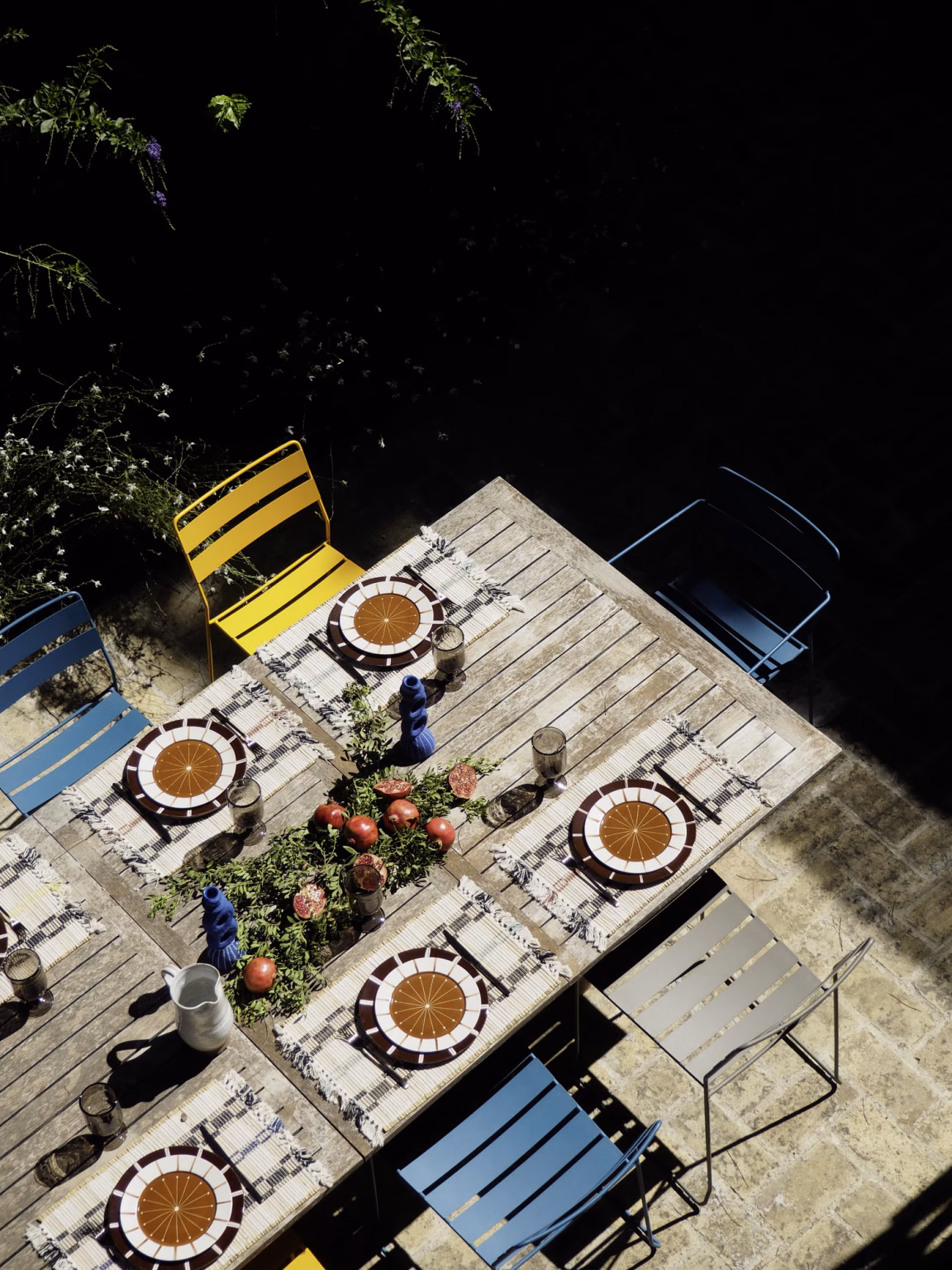 Vista cenital de una mesa de madera exterior puesta con platos, granadas y sillas de colores en Villa Arena