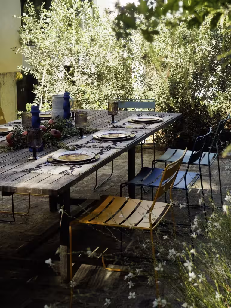 Mesa larga de madera bajo los olivos con sillas azules y amarillas y flores silvestres en Villa Arena