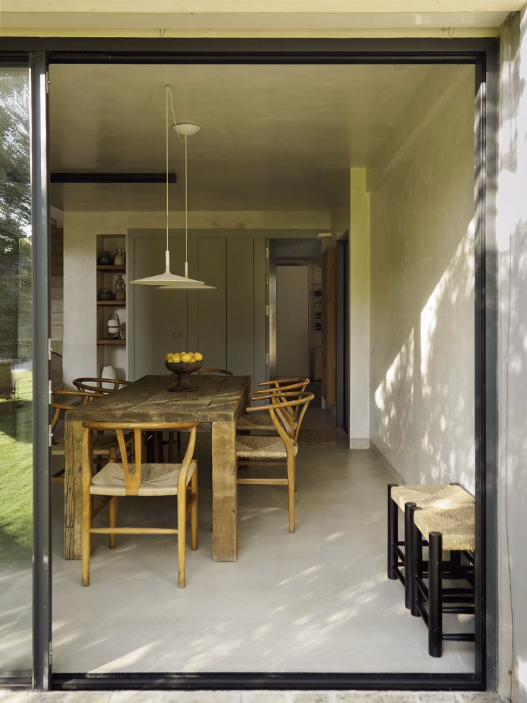 Mesa de comedor de madera con sillas de rejilla y lámpara colgante tras el marco negro de la cristalera en Villa Arena