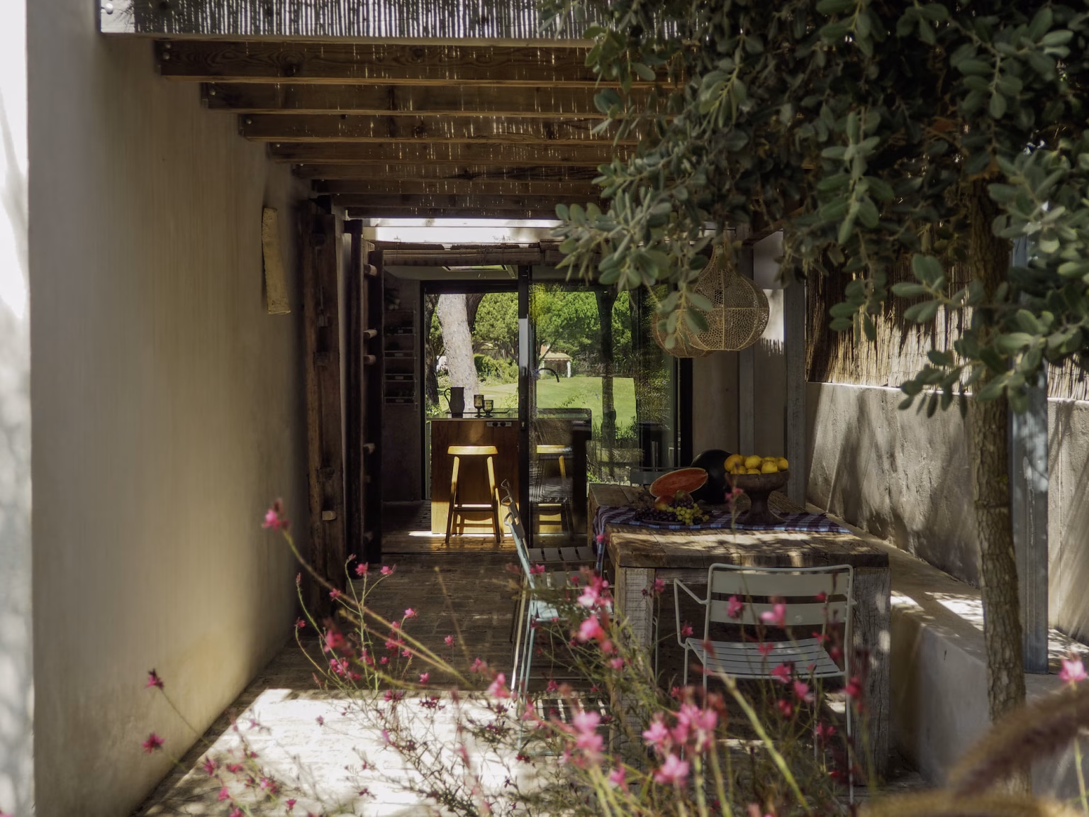 Galería cubierta de vigas y cañizo que atraviesa el jardín, con la mesa exterior al fondo en Villa Arena