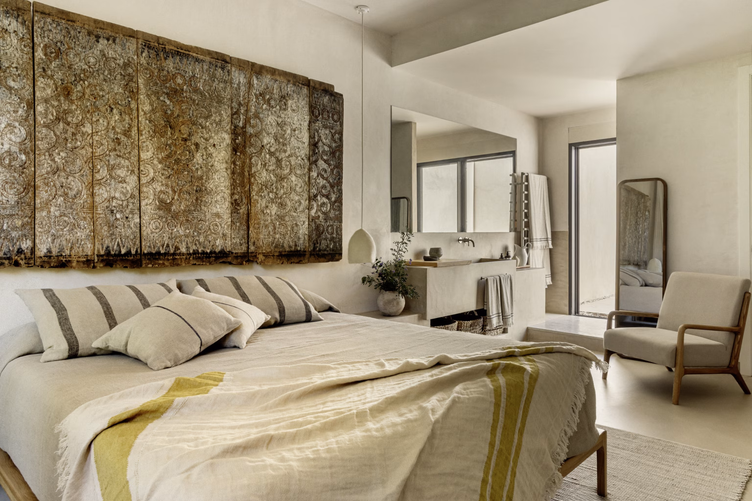 Cama con ropa de lino a rayas bajo un gran panel vegetal y baño en suite al fondo en Villa Arena de ÁBATON