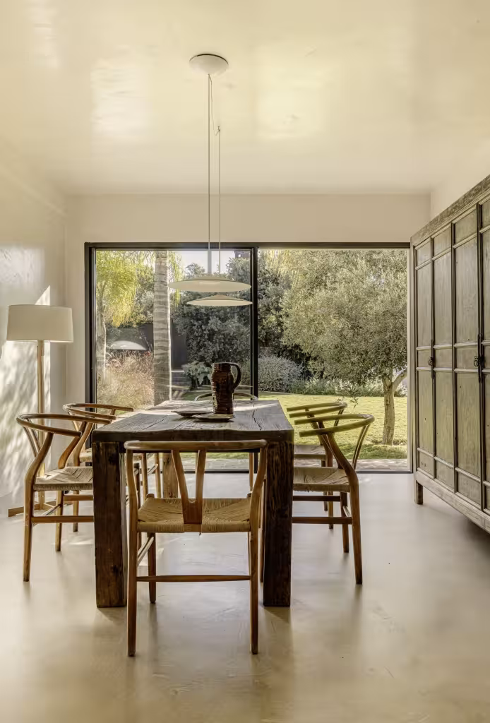 Comedor de mesa de madera maciza y sillas de rejilla ante una cristalera al jardín de Villa Arena