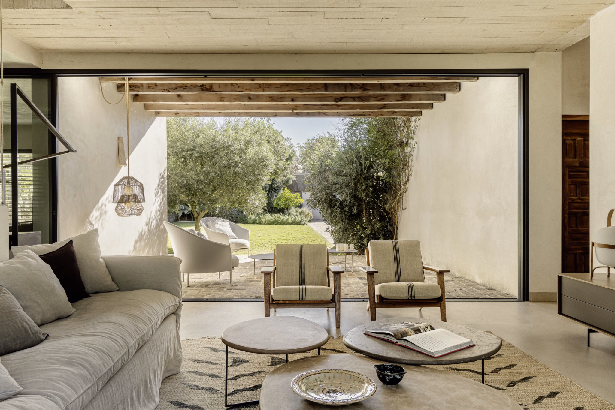 Salón de Villa Arena abierto al jardín con olivos a través de un gran hueco bajo vigas de madera