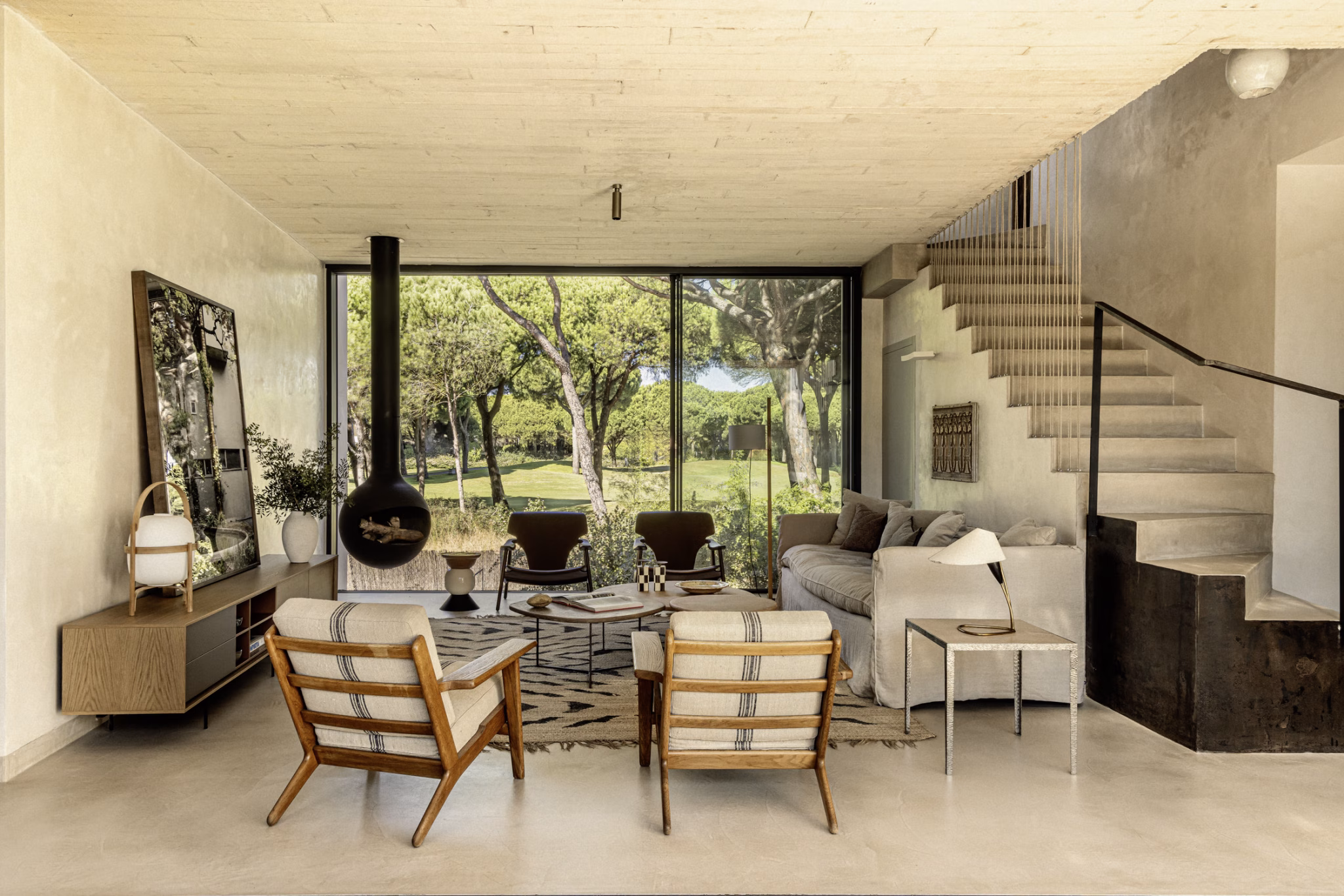 Salón de Villa Arena con chimenea suspendida, butacas de madera y escalera blanca ante el jardín