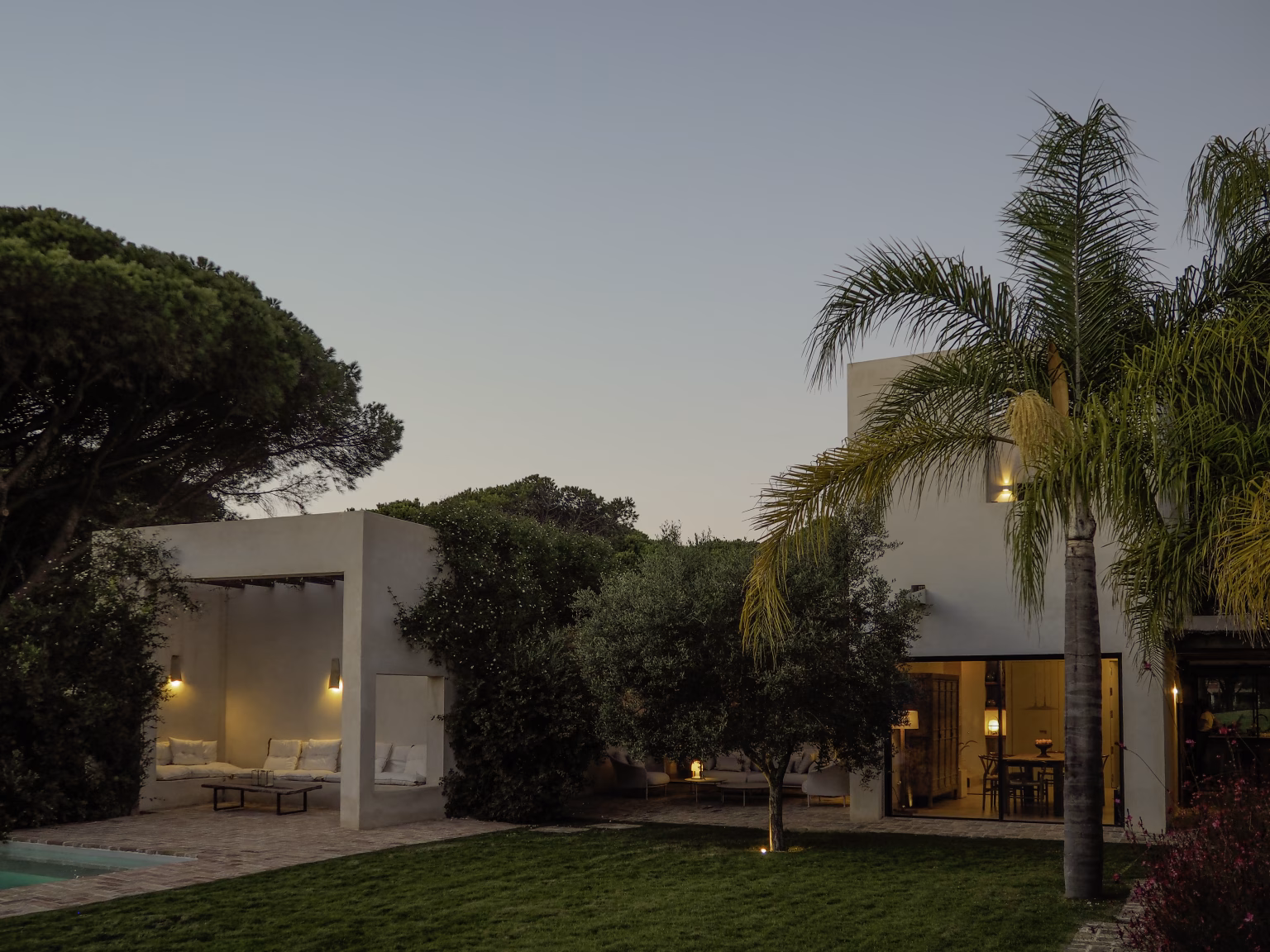Jardín de césped y palmera ante la fachada blanca de Villa Arena al atardecer