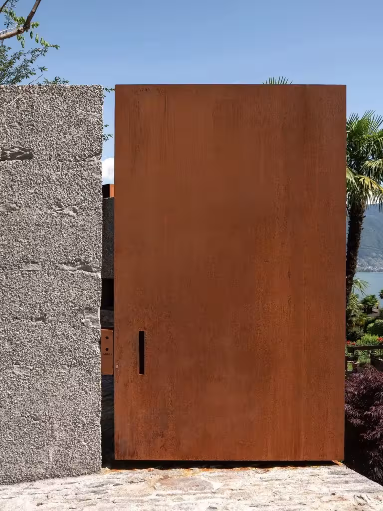 Puerta pivotante de acero corten encajada en el muro de piedra de la casa San Nazzaro