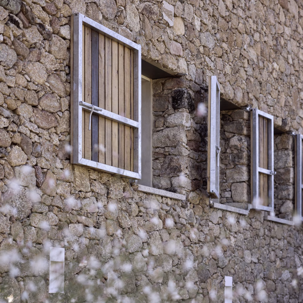 Muro de mampostería con contraventanas de madera y marco blanco en la fachada de la casa TLV