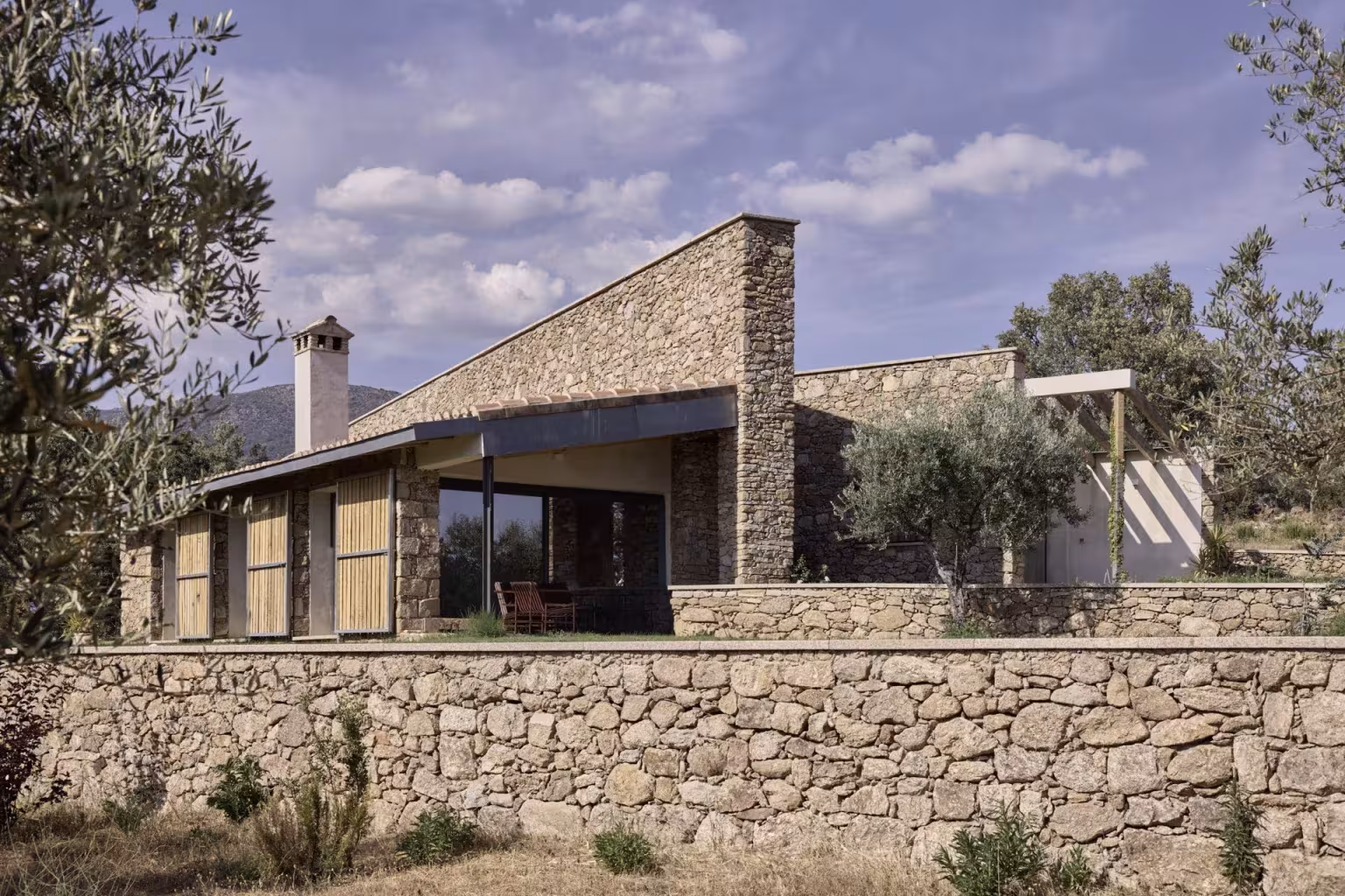 Casa de campo del Proyecto TLV de Ábaton con muro de piedra, cubierta a dos aguas y olivos en primer plano