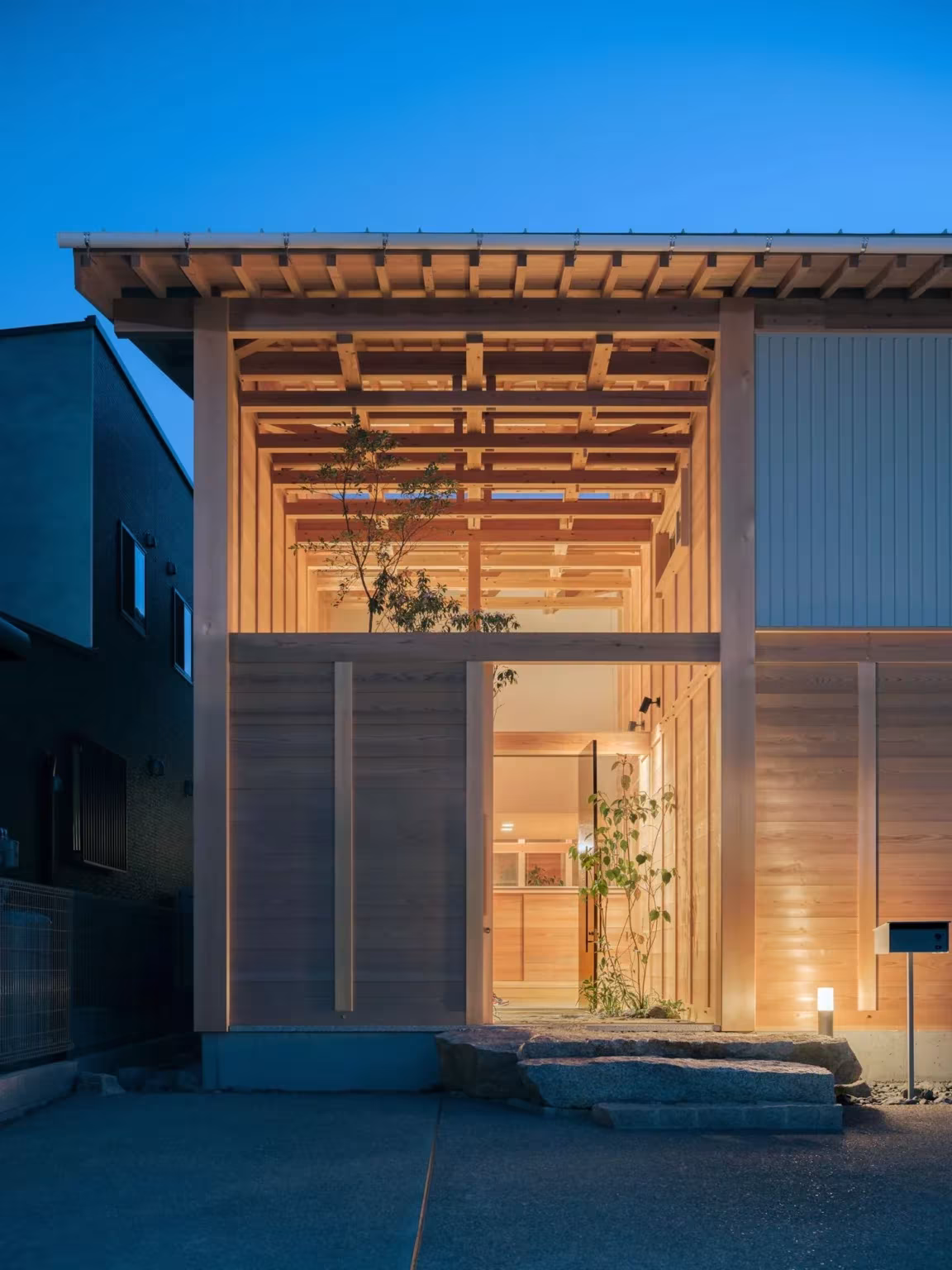 Casa en Aichi al anochecer con el enrejado de cedro iluminado desde el interior y la entrada con luz cálida