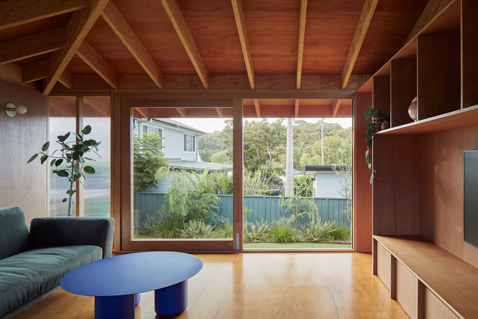 Sala de Princes Studio con sofá verde, mesa azul y ventanal al jardín bajo cubierta de vigas de madera, Curious Practice